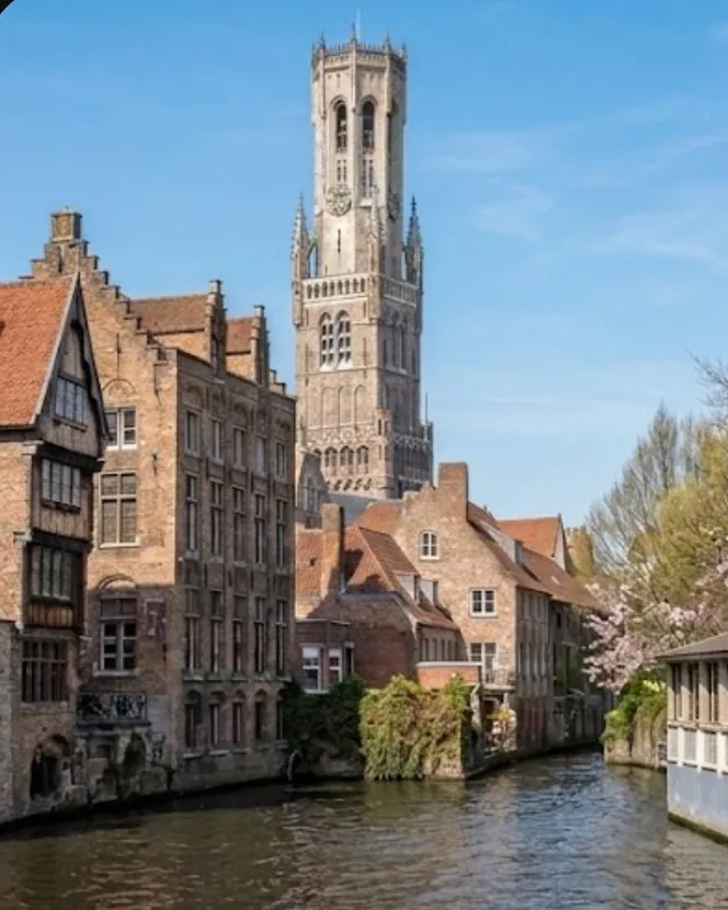 The same corner of Bruges in spring: a pale blue sky, a cherry tree in blossom to the right and the canal lying still.
