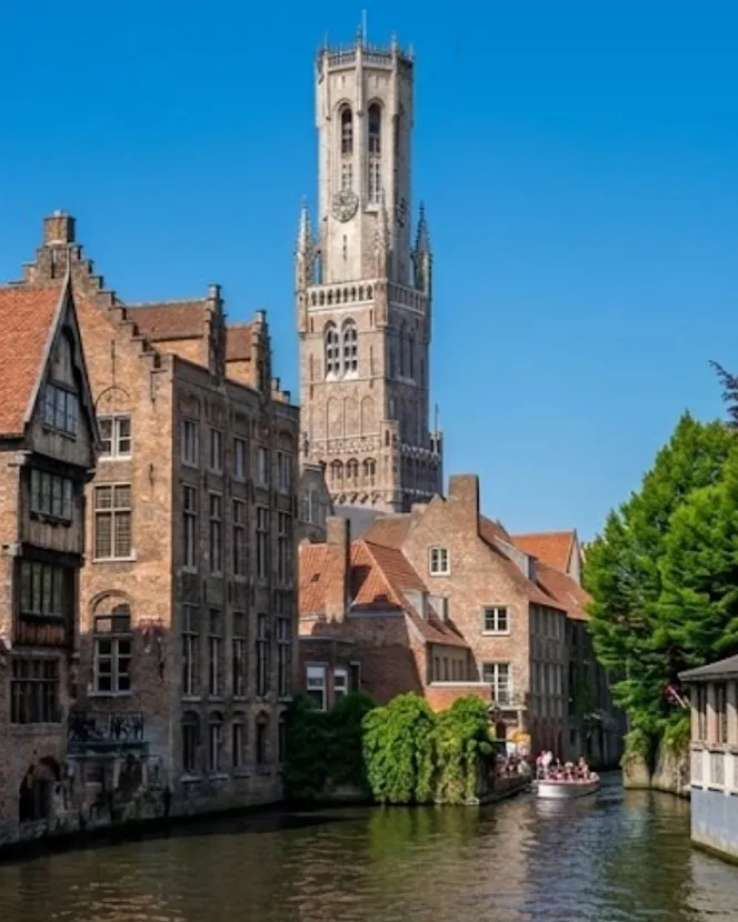 The same corner of Bruges in summer: a deep blue sky, the trees in full leaf and a boat of visitors crossing the canal.