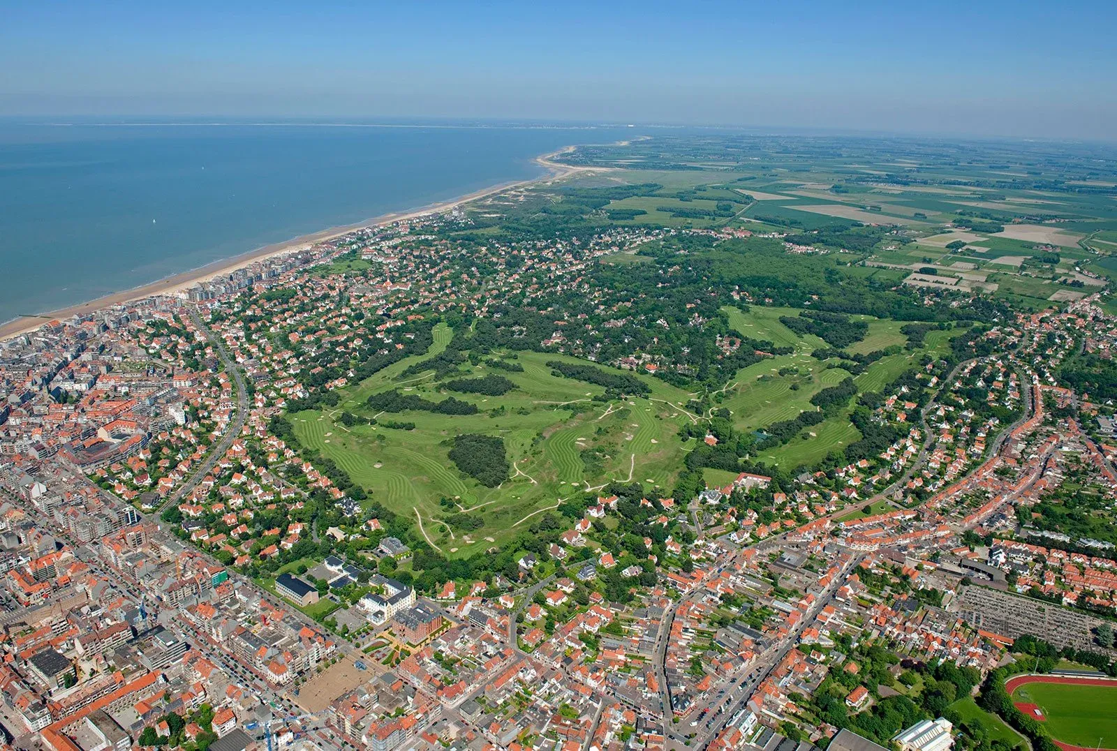 Knokke from the air: the North Sea and its beach along one edge, the red roofs of the town all around, the green of the golf course at its centre, and the polders reaching inland.